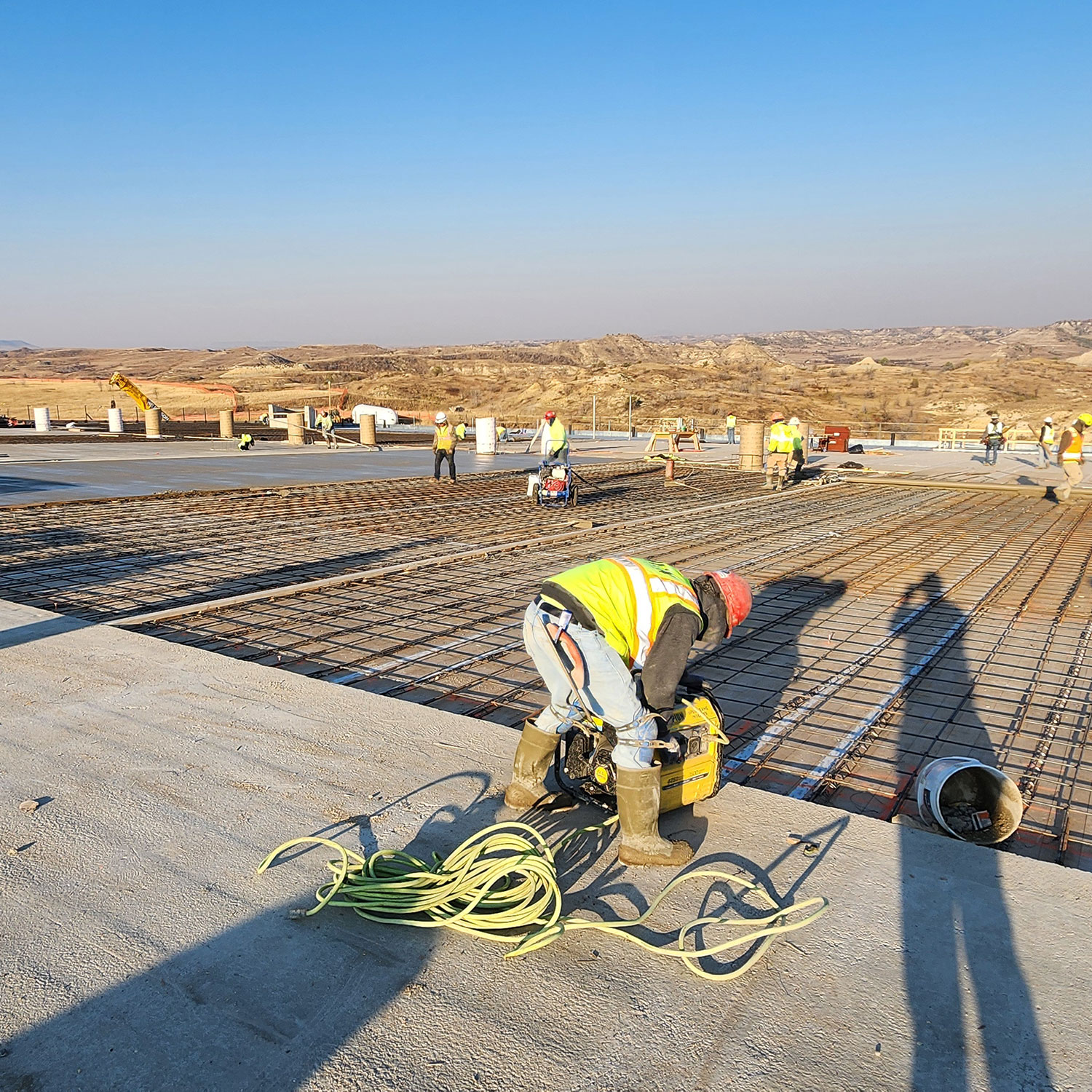 Construction workers in safety vests and hard hats prepare and install rebar for a concrete slab on a large construction site, surrounded by hilly terrain under a clear blue sky