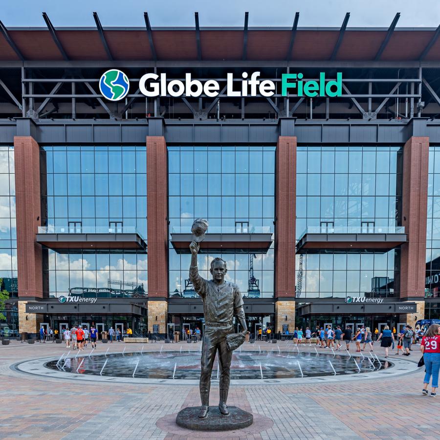 Statue of a man holding a baseball helmet above his head in one hand and a baseball glove to his side in the other hand with a building in the background